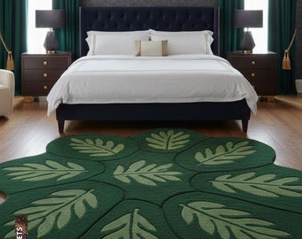May include: A green leaf-shaped rug with a detailed leaf pattern in a bedroom setting. The rug is placed in front of a bed with white bedding and a dark blue headboard. The room has dark green curtains and matching nightstands.