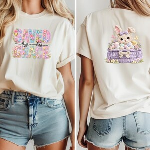 May include: Cream-coloured t-shirt with the text "SAVED by His GRACE" in a floral design. The back of the shirt features a bunny in a basket with Easter eggs and flowers. The design is in pastel colours.
