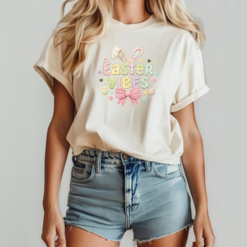 May include: A cream-coloured t-shirt featuring a colourful Easter graphic. The graphic includes the text "Easter Vibes", bunny ears, a bow, and Easter eggs. The shirt is worn with denim shorts.
