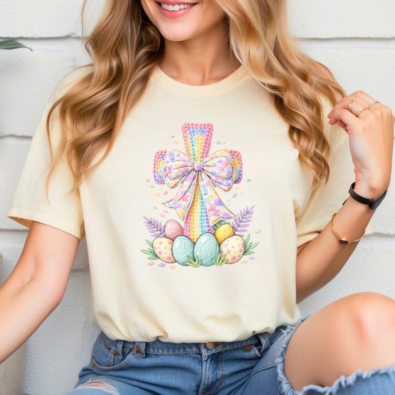 May include: A pale yellow t-shirt with a colourful Easter cross design. The cross is decorated with a large bow and surrounded by decorated Easter eggs and greenery. The design uses pastel colours.