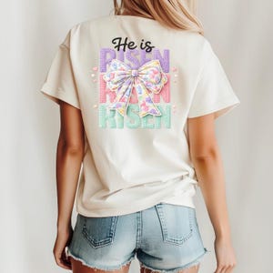 May include: A cream-coloured t-shirt featuring the text "He is Risen" in a layered design. The design includes a large bow with pastel floral patterns. The shirt is worn with denim shorts, creating a casual, spring-themed look.
