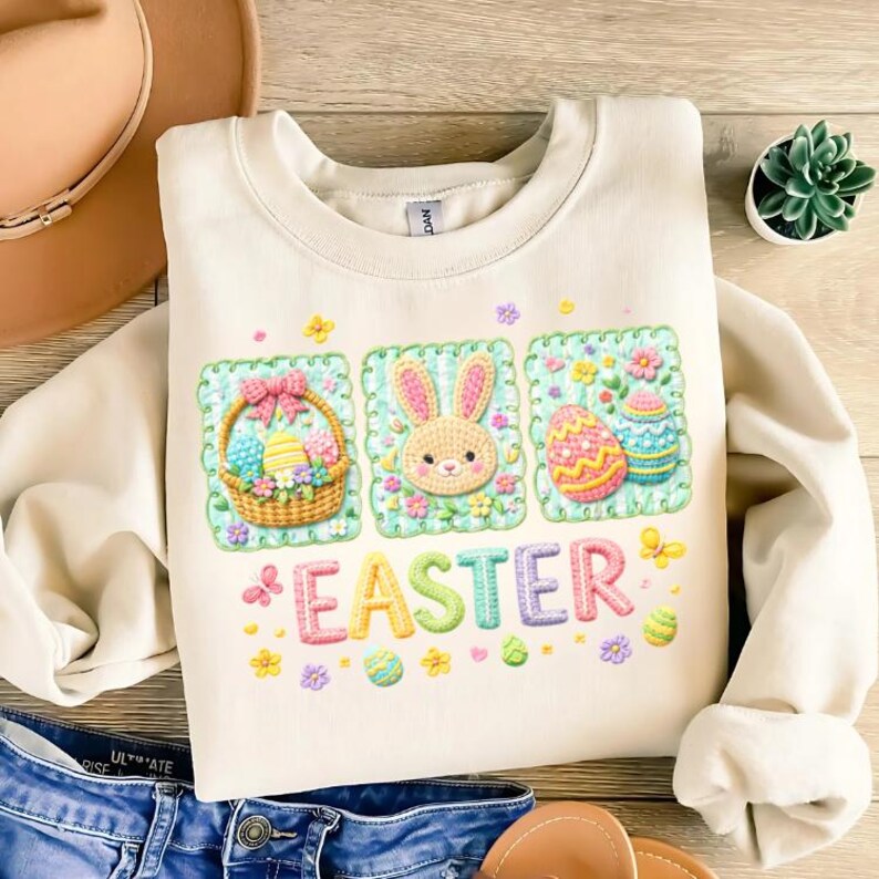 May include: Cream-coloured sweatshirt featuring an Easter design. The design includes a basket of eggs, a bunny face, and decorated eggs, with the word "EASTER" in colourful letters. The sweatshirt is made of a soft material.