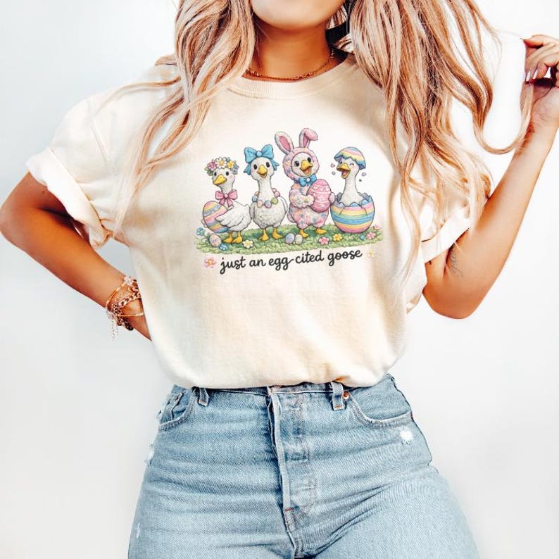 May include: A cream-coloured t-shirt with an Easter graphic. The design shows four cartoon geese in Easter costumes, with the text "just an egg-cited goose" underneath. The geese are standing on a patch of green grass.