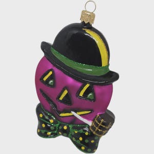 Può includere: Un ornamento a tema Halloween a forma di zucca viola con un cappello nero e una striscia verde e gialla. La zucca ha una faccia intagliata con accenti gialli e un papillon nero e verde a pois. Sta tenendo una pipa.