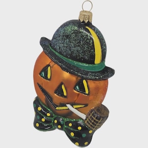 Peut inclure: Ornement d'Halloween en forme de citrouille avec un chapeau noir, une bordure verte et une rayure jaune. La citrouille a un visage sculpté et fume une pipe. Un nœud papillon noir à pois jaunes complète le look festif.
