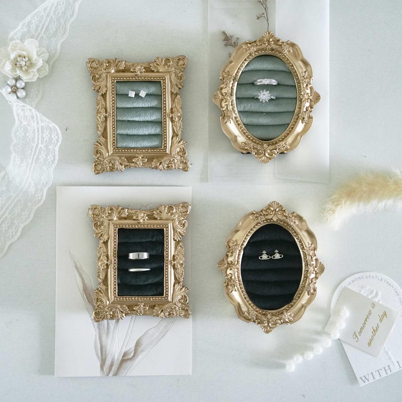 May include: Four ornate gold-toned frames used as jewelry displays. Two rectangular frames contain sage green and black velvet ring slots. Two oval frames also feature sage green and black velvet ring slots. Various silver rings are placed in the slots.