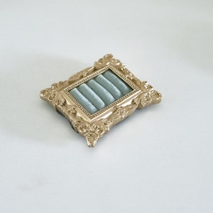 May include: A rectangular jewelry display tray featuring an ornate gold-colored frame with intricate scrollwork detailing. The interior of the tray is lined with soft, light blue velvet fabric, which is molded into four parallel horizontal slots.