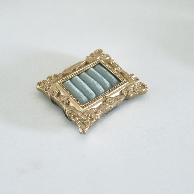 May include: A rectangular jewelry display tray featuring an ornate gold-colored frame with intricate scrollwork detailing. The interior of the tray is lined with soft, light blue velvet fabric, which is molded into four parallel horizontal slots.