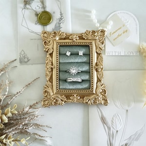 May include: A rectangular gold-toned ornate frame with a sage green velvet insert featuring five horizontal slots. The display holds two square stud earrings, a round halo-style ring, and a silver-toned band ring with a central stone setting.