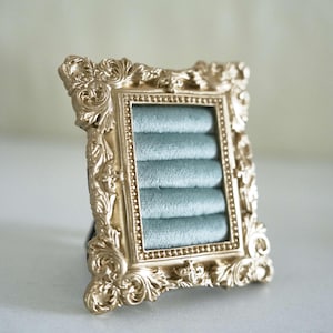 May include: A rectangular jewelry display stand featuring an ornate, metallic gold frame with intricate scrollwork and a beaded inner border. The center contains five horizontal rows of soft, light blue velvet padding designed to hold rings.