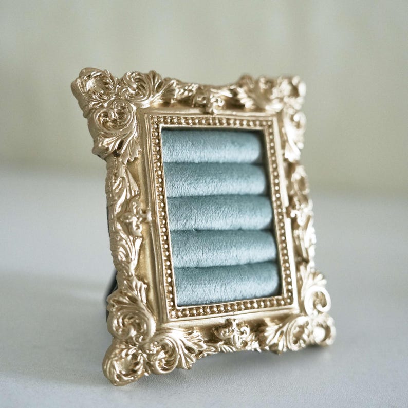 May include: A rectangular jewelry display stand featuring an ornate, metallic gold frame with intricate scrollwork and a beaded inner border. The center contains five horizontal rows of soft, light blue velvet padding designed to hold rings.