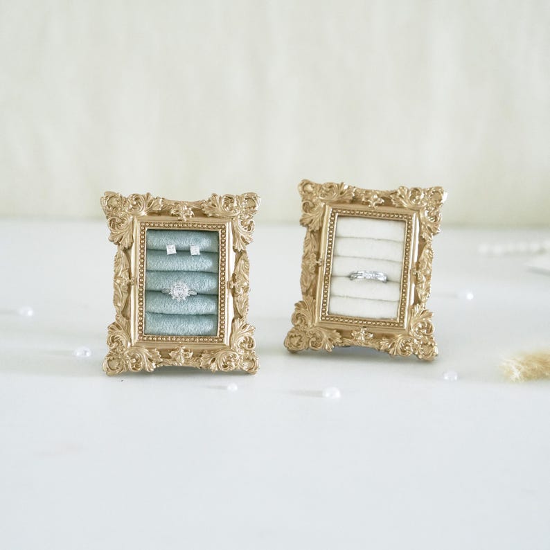 May include: Two rectangular jewelry display stands with ornate gold frames. The left stand features a light blue velvet insert holding two small stud earrings and one ring. The right stand features a white velvet insert holding one silver ring.