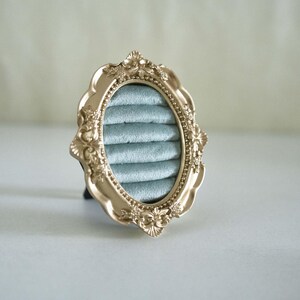 May include: An oval ring display holder featuring a metallic gold frame with ornate, raised floral and scrollwork detailing. The interior is lined with soft, light blue velvet fabric arranged in five horizontal padded rows for holding rings.