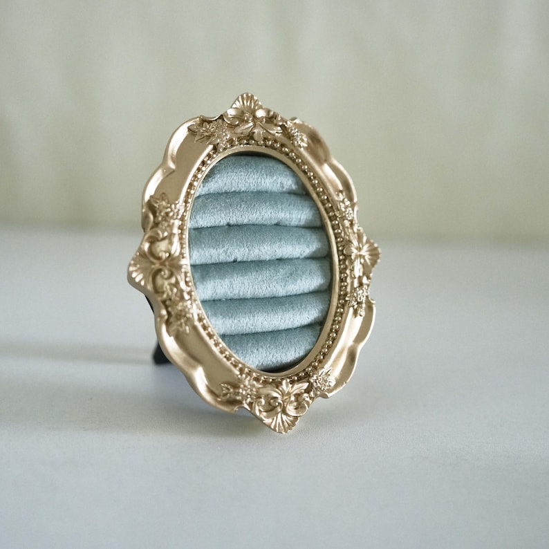 May include: An oval ring display holder featuring a metallic gold frame with ornate, raised floral and scrollwork detailing. The interior is lined with soft, light blue velvet fabric arranged in five horizontal padded rows for holding rings.