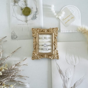 May include: A rectangular gold-toned display frame with ornate, scroll-like detailing along the edges. The interior features a cream-colored velvet insert with four horizontal slots, currently holding two small stud earrings and two silver-toned rings.