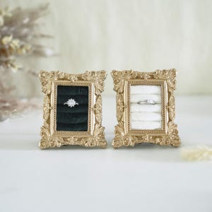 May include: Two rectangular gold-toned frames with ornate, scroll-like detailing. Each frame contains a velvet ring insert, one in black and one in white. A silver-toned ring with a central stone sits in the black insert, and a plain band in the white.