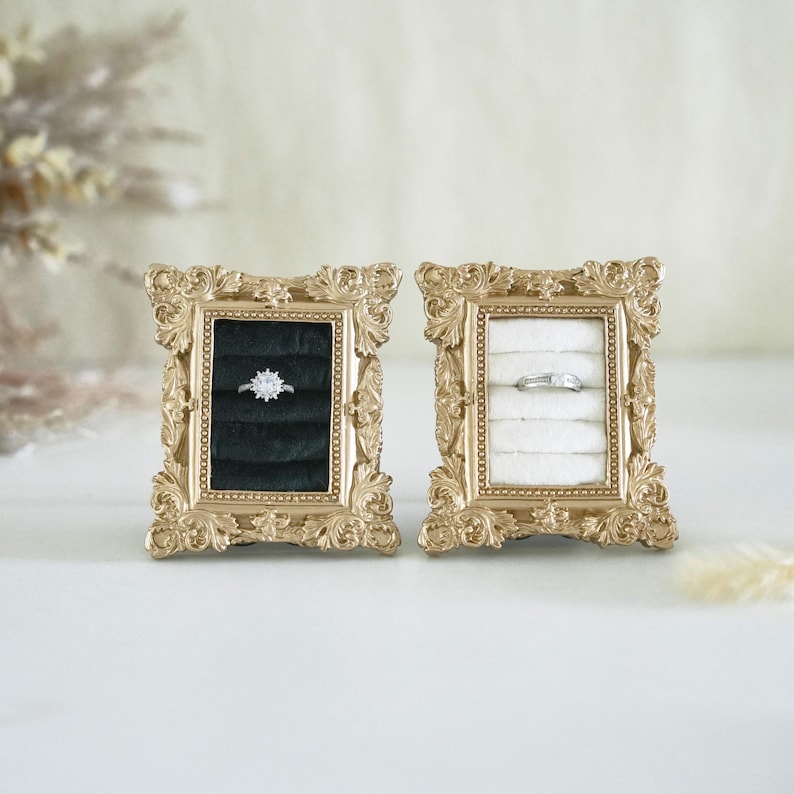May include: Two rectangular gold-toned frames with ornate, scroll-like detailing. Each frame contains a velvet ring insert, one in black and one in white. A silver-toned ring with a central stone sits in the black insert, and a plain band in the white.