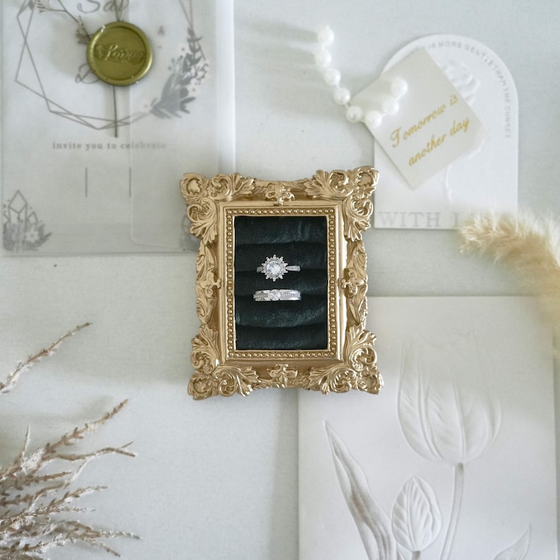 May include: A rectangular gold-toned ornate frame with a black velvet insert holding two silver-toned rings. The top ring features a starburst design with a central stone, while the bottom ring has a rectangular stone. The frame is surrounded by stationery.
