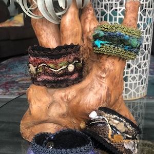 May include: Four handmade fabric bracelets with different colors and textures. The bracelets are displayed on a wooden branch with a white lantern in the background.