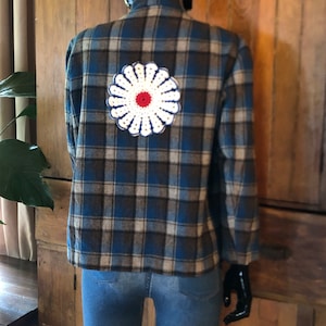 May include: A blue and brown plaid shirt with a white crocheted flower on the back. The flower has a red center.