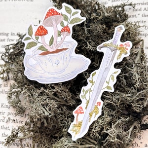 Może przedstawiać: Dwie naklejki o tematyce fantasy. Jedna przedstawia grzyby wyrastające z filiżanki, druga miecz z liśćmi i grzybami. Naklejki to ilustracje akwarelowe. Tło jest porośnięte mchem.