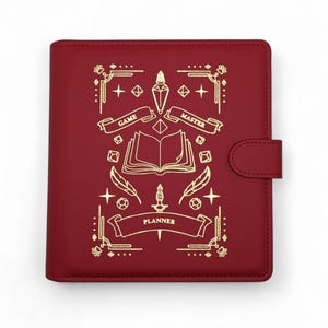 May include: A burgundy planner with gold foil accents. The cover features an open book, feathers, and the words "Game Master Planner." A tab closure is on the right side. The design is reminiscent of fantasy role-playing games.