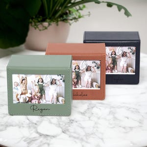 May include: Three personalized photo boxes in sage green, brown, and black. Each box displays a framed photo of children and a name. The boxes are square and made of a leather-like material. A plant in a pot is in the background.