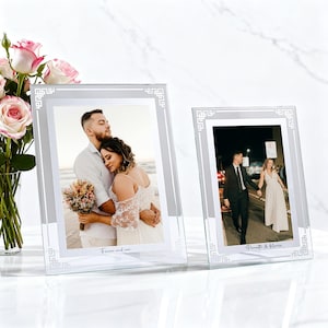 Puede incluir: Dos marcos de fotos de vidrio transparente con bordes plateados y diseños decorativos en las esquinas. Cada marco contiene una fotografía de una pareja. Los marcos están sobre una superficie blanca, con un ramo de rosas rosas en el fondo. El texto "Forever and ever" es visible.