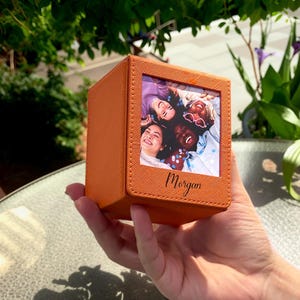 May include: An orange photo box with a picture of four people smiling. The box has a textured surface and the name "Morgan" is written in cursive on the front.