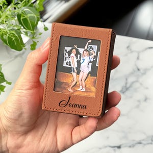 May include: A brown leather wallet with a photo of two people. The wallet has a rectangular window for the photo and the name "Joanna" is written in cursive below the photo. The wallet is held in a hand.
