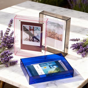 May include: Three acrylic photo frames in pink, gray, and blue. The pink frame holds a photo of a bridge, the gray frame a beach scene, and the blue frame a tablet. Lavender sprigs are arranged around the frames.