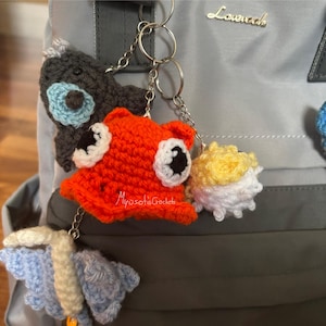 May include: A collection of handmade crochet keychains in various shapes and colors. The keychains include a gray whale, an orange fish, a yellow and white creature, and a blue butterfly. Each keychain is attached to a silver keyring.