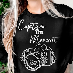 May include: Black sweatshirt with the phrase "Capture The Moment" in white script above a white line drawing of a camera. The sweatshirt is a crew neck style.