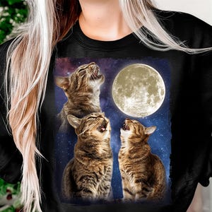 May include: Black sweatshirt featuring a graphic of three tabby cats howling at a full moon against a starry night sky. The cats are brown and tan, with open mouths. The moon is a bright yellow circle.
