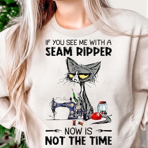 May include: A beige sweatshirt featuring a grumpy grey cat with yellow eyes, a sewing machine, and the text "IF YOU SEE ME WITH A SEAM RIPPER NOW IS NOT THE TIME."