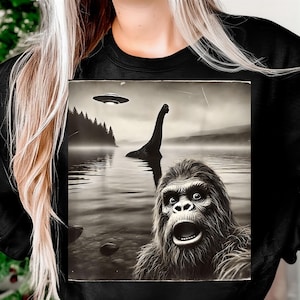 May include: Black sweatshirt featuring a grayscale graphic of a lake scene with a UFO, a long-necked creature, and a surprised ape-like creature. The image has a vintage, slightly grainy aesthetic.