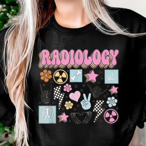 Könnte beinhalten: Schwarzes Sweatshirt mit dem Wort "RADIOLOGY" in rosa Retro-Schrift. Das Design umfasst verschiedene medizinische Grafiken wie einen Knochen, ein Strahlungssymbol, eine Skeletthand und Blumen.