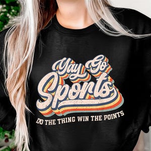 Op de afbeelding: Zwart sweatshirt met de tekst "Yay Go Sports" in een retro lettertype met blauwe, oranje en gele strepen. De zin "Do the thing win the points" staat eronder.