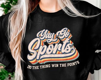 Op de afbeelding: Zwart sweatshirt met de tekst "Yay Go Sports" in een retro lettertype met blauwe, oranje en gele strepen. De zin "Do the thing win the points" staat eronder.
