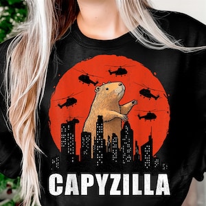 May include: Black sweatshirt featuring a cartoon capybara with helicopters and city skyline against a red sun. The word "CAPYZILLA" is printed in white block letters at the bottom of the design.