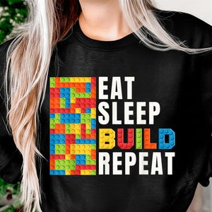 May include: Black sweatshirt featuring the text "EAT SLEEP BUILD REPEAT" in white, with the word "BUILD" in colourful block letters. A colourful block design is on the left side of the text.