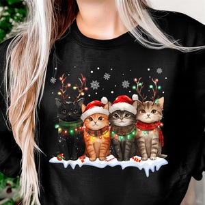 Peut inclure: Sweat-shirt noir avec quatre chats décorés pour Noël. Chaque chat porte un bonnet de Père Noël, une écharpe et des guirlandes lumineuses. Un chat a des bois lumineux. Des flocons de neige et de la neige complètent le motif.