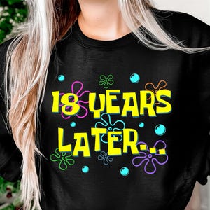 May include: Black sweatshirt with the text "18 YEARS LATER..." in yellow, with colorful cartoon-style accents. The design includes pink, green, and blue floral and bubble graphics. A fun, casual top.