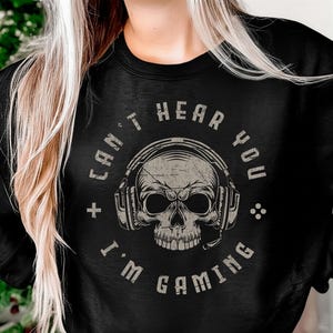 Op de afbeelding: Zwart sweatshirt met een verweerde afbeelding van een schedel met een koptelefoon. De tekst rond de schedel luidt "CAN'T HEAR YOU I'M GAMING". Het ontwerp is lichtgrijs.