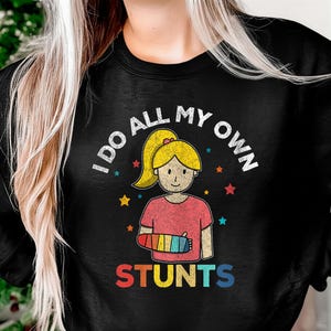 Puede incluir: Sudadera negra con una ilustración de dibujos animados de una niña con un yeso en el brazo. El texto "I DO ALL MY OWN" está arqueado sobre la niña, y "STUNTS" está escrito debajo. La niña tiene el pelo rubio y lleva una camiseta rosa.