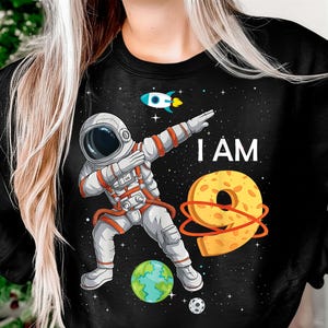 Może przedstawiać: Czarna bluza z kapturem z kreskówkowym astronautą wykonującym dab, z napisem "I AM 9". Projekt zawiera rakietę, planetę Ziemię, żółtą cyfrę 9 i małą piłkę nożną, wszystko na tle gwiezdnego nieba.