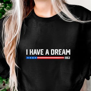 Può includere: Felpa nera con la scritta bianca "I HAVE A DREAM". Sotto, una striscia orizzontale con stelle blu e strisce rosse e bianche, con l'anno "1963" sotto.