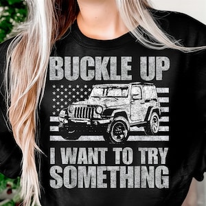 May include: Black sweatshirt with a distressed white graphic. The graphic features a Jeep in front of an American flag design, with the words "BUCKLE UP" above and "I WANT TO TRY SOMETHING" below the vehicle.