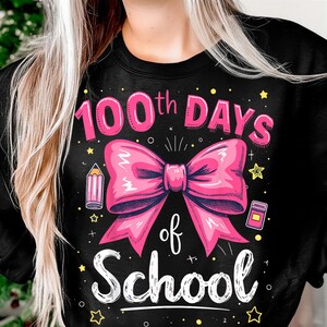 以下が含まれることがあります： ピンクのリボンと「100th Days of School」の文字が入った黒いスウェットシャツ。デザインにはピンクの鉛筆、小さな黄色とピンクの本、黄色の星が含まれています。テキストとグラフィックは遊び心のある手書き風です。