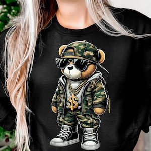 May include: Black sweatshirt featuring a cartoon teddy bear wearing a camouflage hat, jacket, and pants. The bear is accessorized with sunglasses, a gold chain, and a dollar sign pendant. The bear is wearing black and white sneakers.